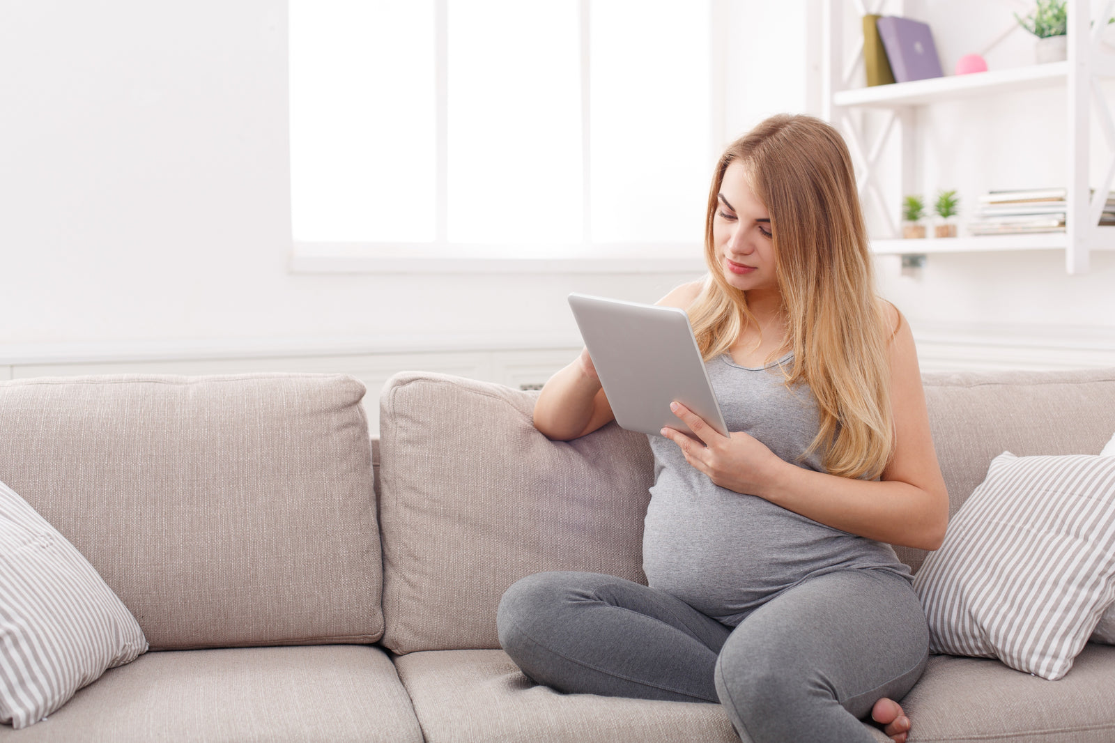 Pregnant woman sat on grey sofa holding a silver tablet