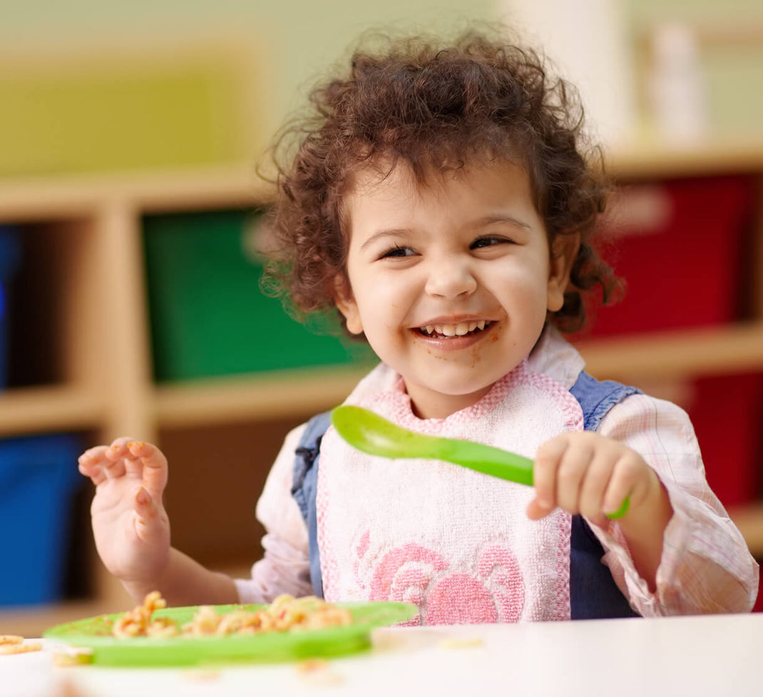 Smiling baby girl with dark curly hair eating her food on a plate with a plastic spoon