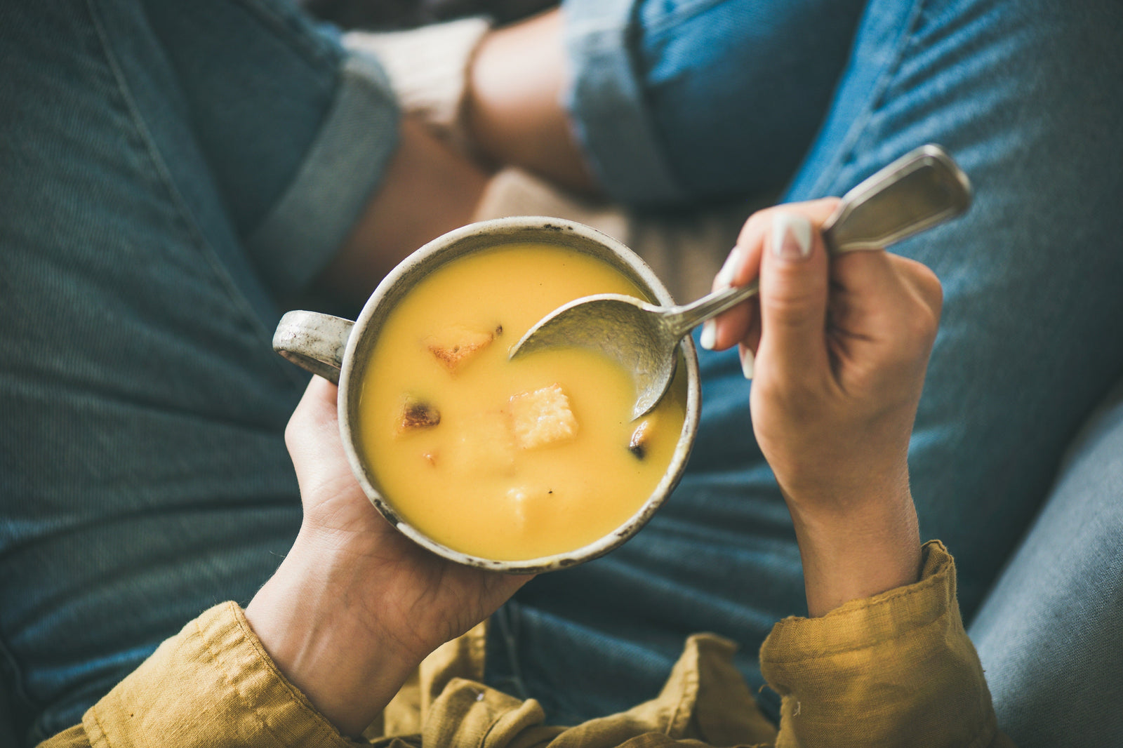Woman with legs crossed eating a bowl of yellow soup with croutons