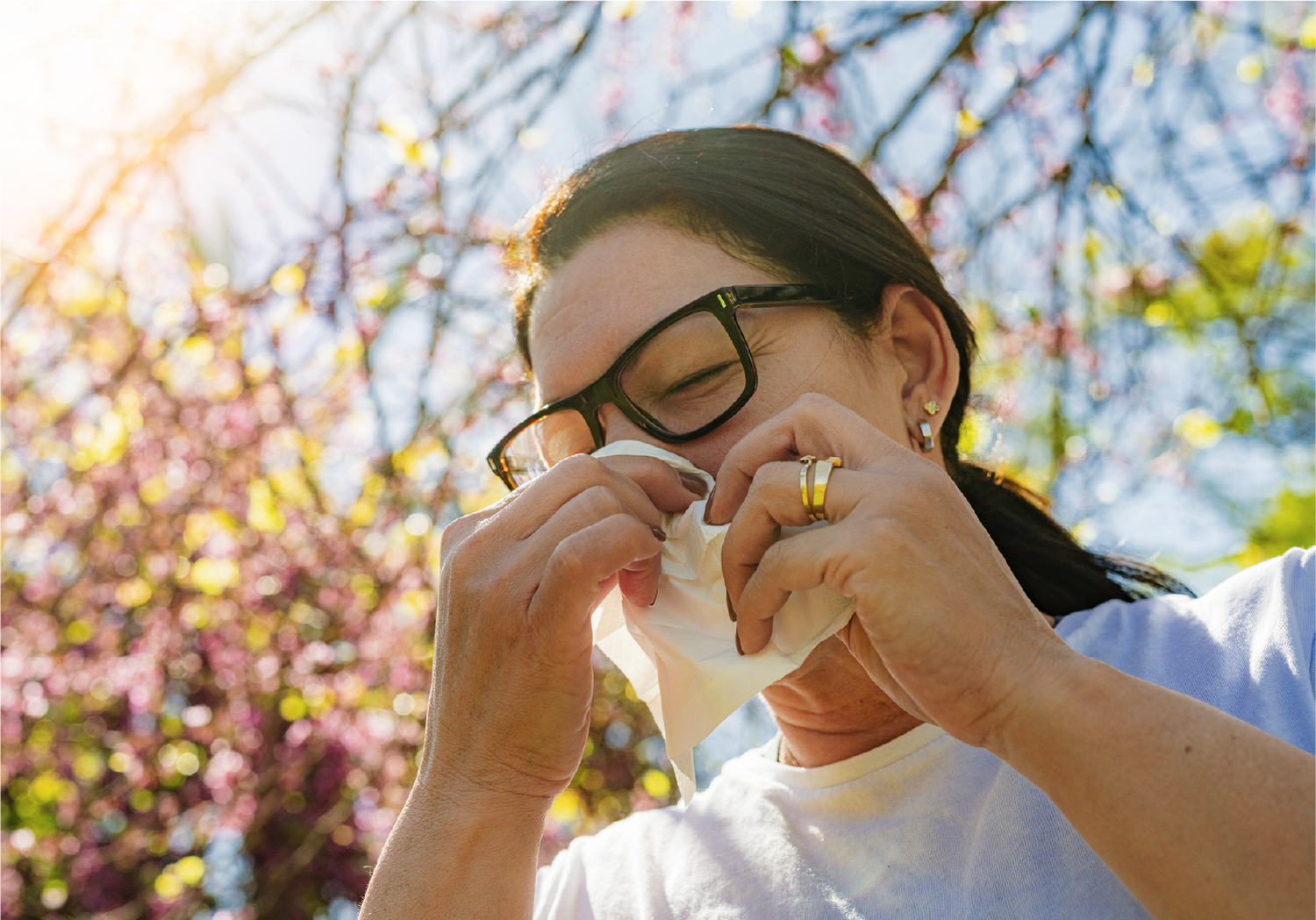 A person sneezing into a tissue outdoors during springtime, with blooming trees and bright sunlight in the background.